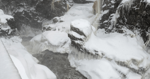 A snowy landscape featuring a frozen waterfall and icy rocks beside a flowing river.