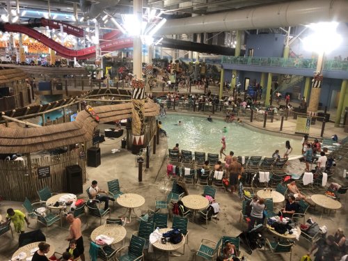 A busy indoor water park with a pool, slides, and many people enjoying the attractions and seating areas.