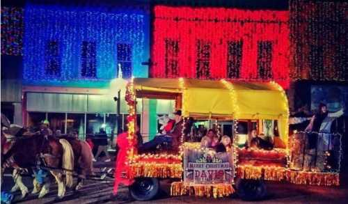 A festive parade float decorated with lights, featuring people celebrating Christmas, with colorful buildings in the background.