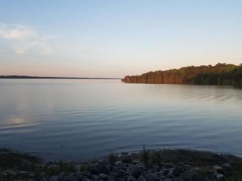 A calm lake at sunset, with gentle ripples and trees lining the shore under a clear sky.