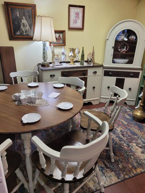 A cozy dining area featuring a round table, white chairs, and vintage decor, including a lamp and glassware.