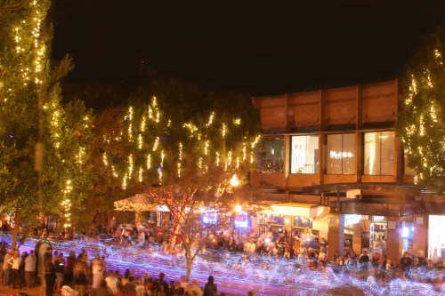 A lively night scene with people gathered outdoors, surrounded by trees adorned with lights and colorful light trails.