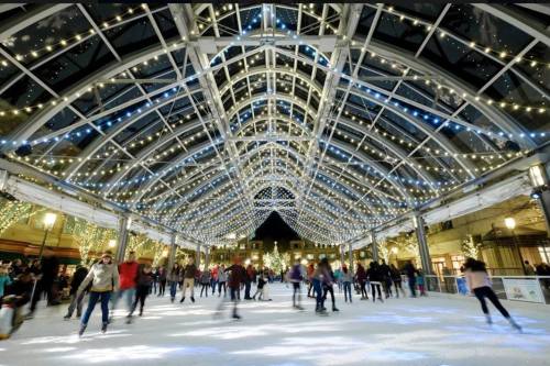 A bustling ice skating rink under a beautifully lit, arched structure with festive lights and people skating.