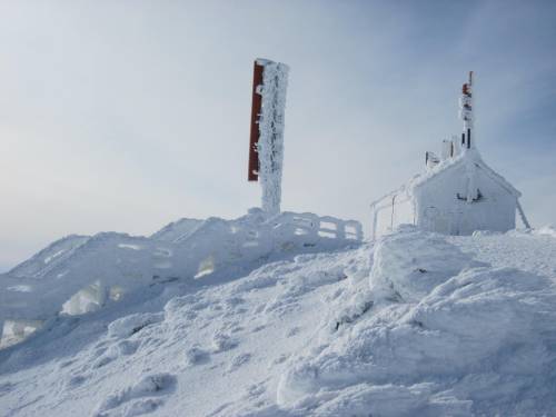A snowy landscape featuring a small building and a tall sign, both covered in thick layers of snow and ice.