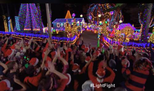 A festive crowd in Santa hats cheers in front of a brightly lit holiday display with colorful lights and decorations.