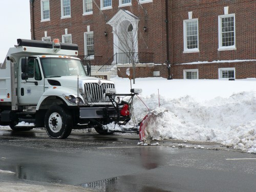 A snowplow truck clearing a snow-covered road in front of a brick building.