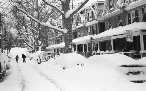 A snowy street lined with houses, covered cars, and two people walking through deep snow.