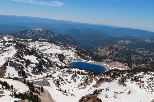 A panoramic view of snow-covered mountains and a blue lake surrounded by forested hills under a clear blue sky.