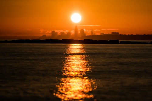 Sunset over a city skyline, with the sun reflecting on calm water and silhouettes of buildings in the background.
