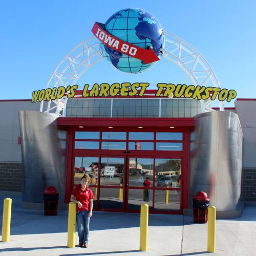 A person stands in front of the Iowa 80 Truckstop entrance, featuring a large globe and "World's Largest Truckstop" sign.