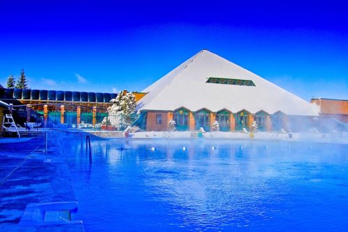 A snowy landscape featuring a pyramid-shaped building beside a steaming blue pool under a clear blue sky.