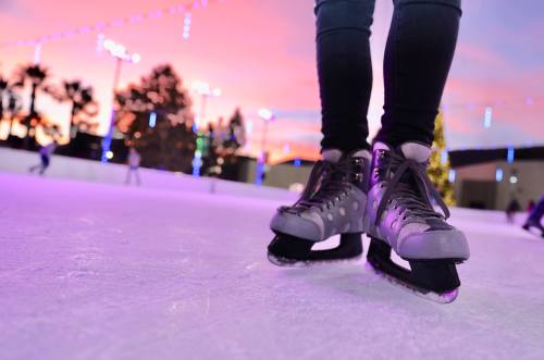 A close-up of ice skates on a rink, with a colorful sunset and festive lights in the background.