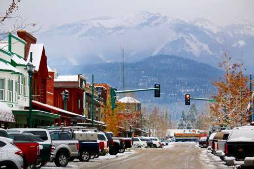 Snow-covered street lined with colorful buildings and parked cars, with mountains in the background under a cloudy sky.