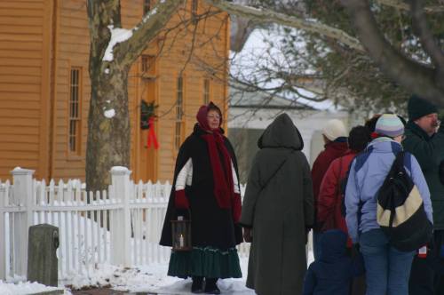 A woman in a historical costume stands near a group of people in winter attire, surrounded by snow and a wooden building.