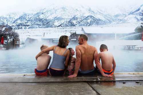 A family of five sits by a steaming hot spring, surrounded by snowy mountains, enjoying a winter day together.