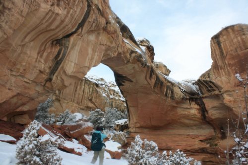 A hiker stands beneath a large rock arch in a snowy canyon, surrounded by red rock formations and sparse vegetation.