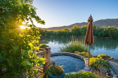 A serene riverside scene at sunset, featuring a hot tub, lush greenery, and a shaded umbrella.