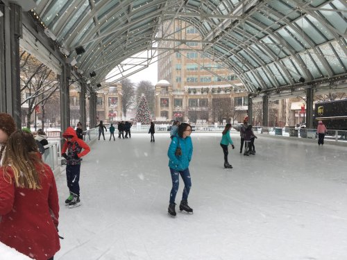 People ice skating under a glass-roofed rink, surrounded by snow and festive decorations in a city setting.