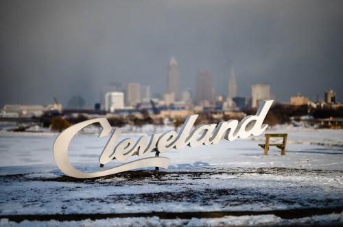 A large "Cleveland" sign in the foreground with a snowy landscape and the city skyline blurred in the background.