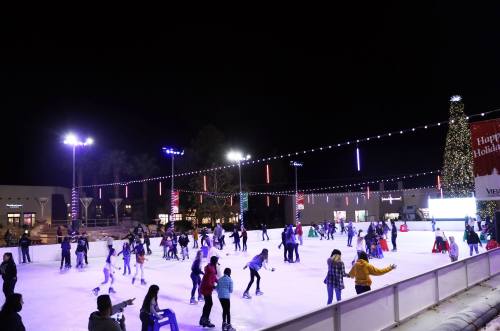 A lively outdoor ice skating rink at night, filled with skaters and festive lights, with a decorated tree in the background.