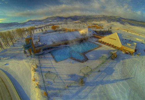 Aerial view of a snowy resort with a steaming pool surrounded by trees and mountains in the background.