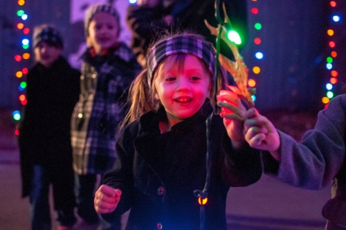 A young girl in a black coat smiles while holding a colorful light, surrounded by festive lights and other children.