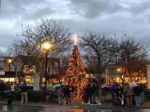 A festive Christmas tree adorned with lights stands in a town square, surrounded by a crowd under a cloudy sky.
