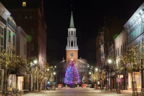 A festive street at night, featuring a large, colorful Christmas tree and a church steeple in the background, adorned with lights.