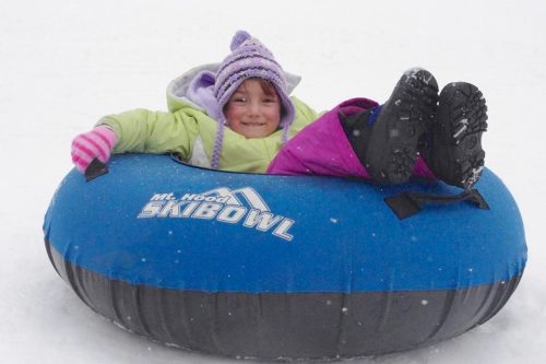 A child in winter clothing smiles while sitting in a blue snow tube, surrounded by falling snow.