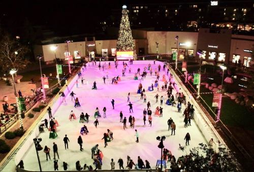 A bustling outdoor ice skating rink at night, decorated with colorful lights and a large Christmas tree in the background.