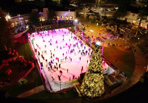 Aerial view of a vibrant ice skating rink surrounded by festive lights and a large decorated Christmas tree.