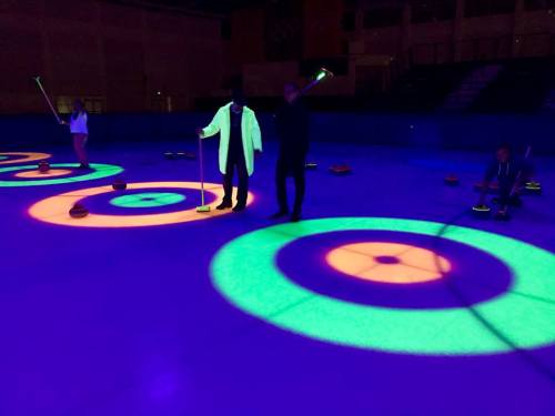 Two people on an illuminated curling rink, holding brooms, with colorful target circles projected on the ice.