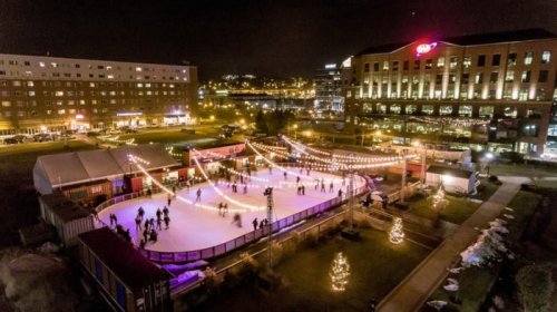 Aerial view of a festive outdoor ice skating rink at night, surrounded by twinkling lights and holiday decorations.