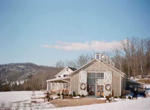 A rustic barn surrounded by snow, with a clear blue sky and hills in the background. Wreaths adorn the barn doors.