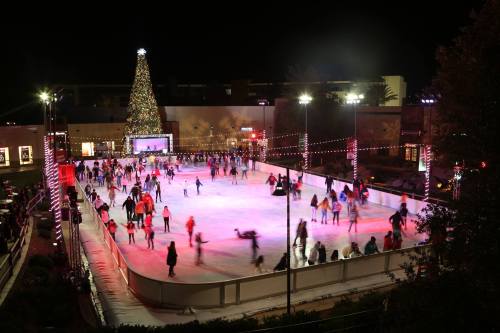 A festive outdoor ice skating rink at night, decorated with lights and a large Christmas tree in the background.
