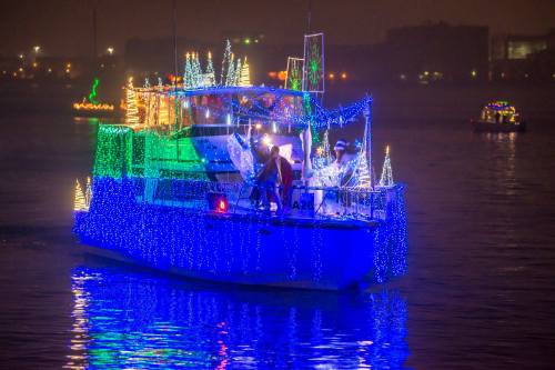 A brightly lit boat decorated with colorful holiday lights floats on water, surrounded by festive decorations.