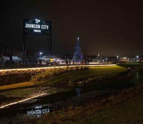 A festive scene in Johnson City at night, featuring a lit Christmas tree and a sign, with reflections in a nearby stream.