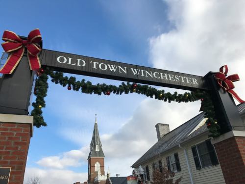 Archway sign reading "Old Town Winchester," decorated with garlands and bows, with a church steeple in the background.