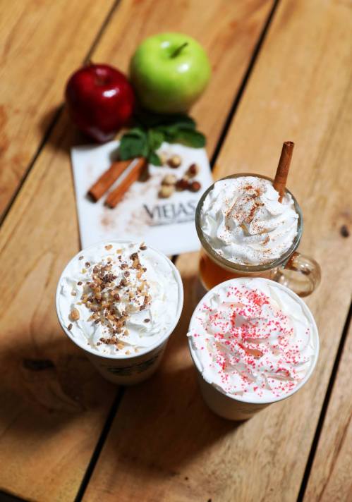 Three festive drinks topped with whipped cream and garnishes, surrounded by apples, cinnamon, and a napkin.