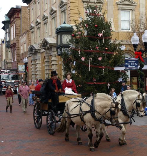 A horse-drawn carriage passes by a decorated Christmas tree in a festive street scene with people enjoying the holiday atmosphere.