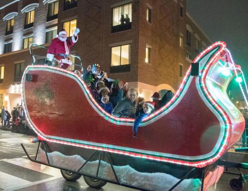 A festive Santa waves from a brightly lit sleigh, surrounded by children, during a nighttime parade.
