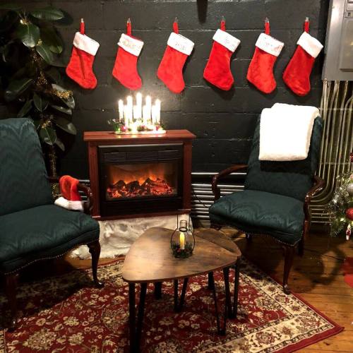 Cozy holiday scene with two chairs, a small table, stockings on the wall, and a fireplace surrounded by festive decor.
