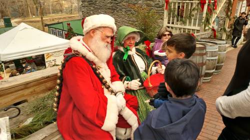 Santa and a woman in green converse with children at a festive outdoor event, surrounded by holiday decorations.