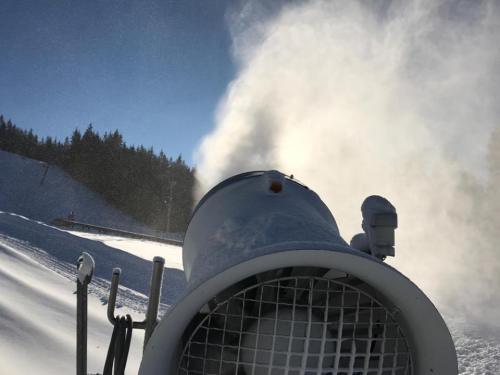 A snowmaking machine sprays artificial snow against a backdrop of trees and a clear blue sky.