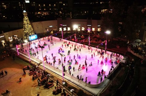 Aerial view of a bustling outdoor ice skating rink, decorated with colorful lights and a Christmas tree nearby.