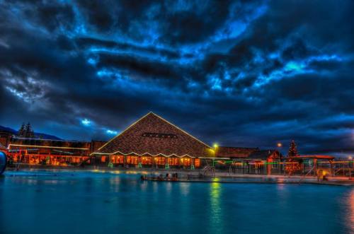 A scenic view of a building by a lake, illuminated with lights under a dramatic, cloudy sky at dusk.