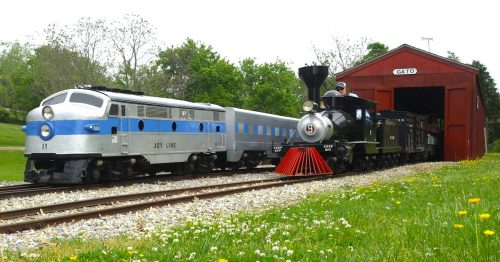 Two model trains, one blue and silver and the other black with a red front, near a red train shed on a sunny day.