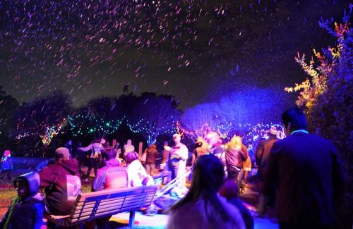 A festive night scene with colorful lights, people enjoying a snowy atmosphere, and a lively outdoor celebration.