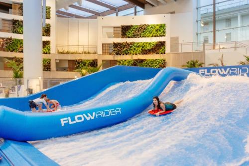 Two people enjoying a FlowRider wave simulator in a modern indoor setting with greenery in the background.