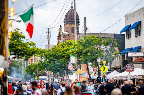 A bustling street festival with crowds, food stalls, and a historic building in the background under a cloudy sky.